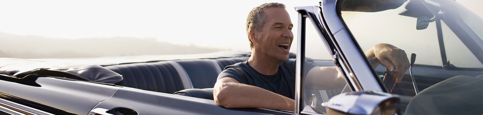 A man with grey hair laughing while driving a convertible