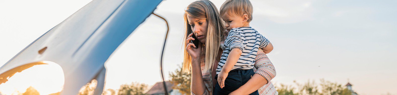 Woman holding toddler on phone next to broken down car